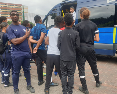 group of young people interacting outside a police van with one person smiling and showing a thumbs up engaging in community activities related to teamwork and youth empowerment