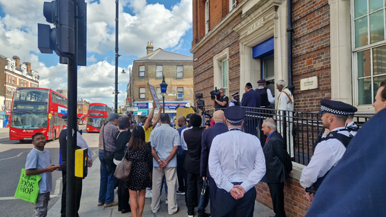 crowd gathered outside building with police presence and media coverage in urban setting two buses passing by under blue sky