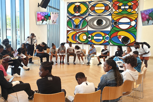 group of young people seated in a circle engaged in an activity in a brightly lit room with colorful artwork on the walls and digital screens